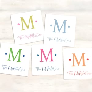 K&ouml;nnte beinhalten: F&uuml;nf wei&szlig;e quadratische Karten mit dem Buchstaben "M" in verschiedenen Farben. Jede Karte tr&auml;gt den Text "The McAllisters" unter dem Buchstaben.