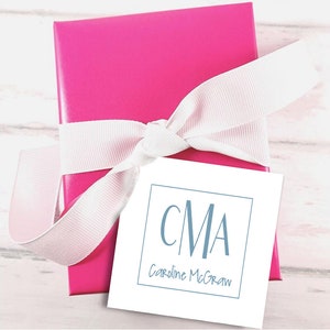 May include: A bright pink gift box tied with a white ribbon. A white tag with the initials "CMA" in blue and the name "Caroline McGraw" is attached to the ribbon. The box is on a light wood surface.