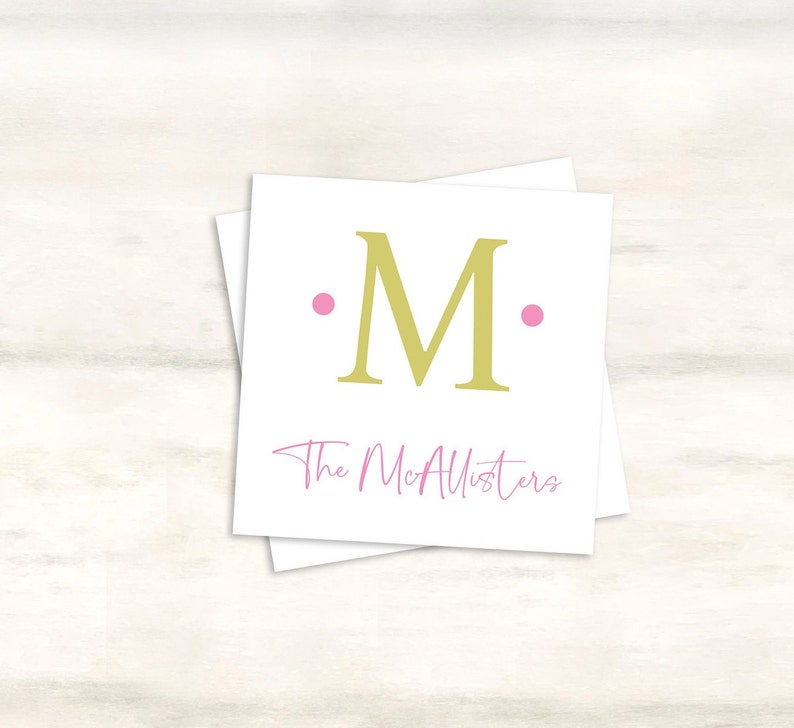 K&ouml;nnte beinhalten: Eine wei&szlig;e Karte mit einem goldenen Buchstaben "M", umgeben von zwei rosa Punkten. Der Text "The McAllisters" ist in rosa Kursivschrift unter dem Buchstaben geschrieben.