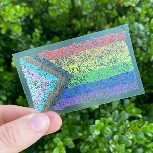 May include: A clear plastic sticker with a rainbow pride flag design made of flowers. The flowers are outlined in different colors of the rainbow.