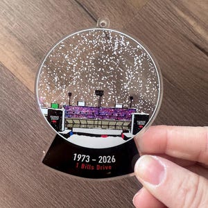May include: A clear plastic snow globe ornament featuring a black and white illustration of a stadium with the text "Highmark Stadium" and "1 Bills Drive" on the base. The snow globe has white glitter inside and the text "1973 - 2026" on the base.