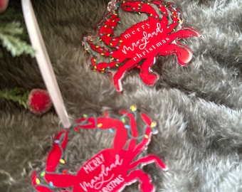 May include: Two red crab-shaped Christmas decorations with white string hangers. The crabs have white lights around them and the text "Merry Maryland Christmas" on them.