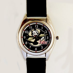 May include: A silver-toned wristwatch with a black leather band. The watch face features a black background with a white cartoon of Felix the Cat. The watch face also has red and white numbers.