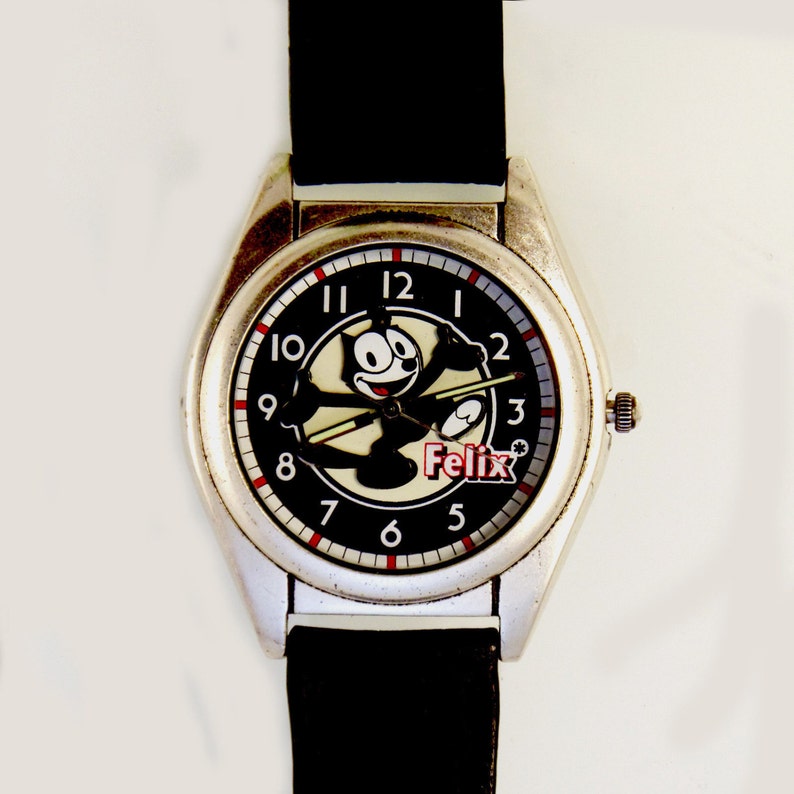 May include: A silver-toned wristwatch with a black leather band. The watch face features a black background with a white cartoon of Felix the Cat. The watch face also has red and white numbers.
