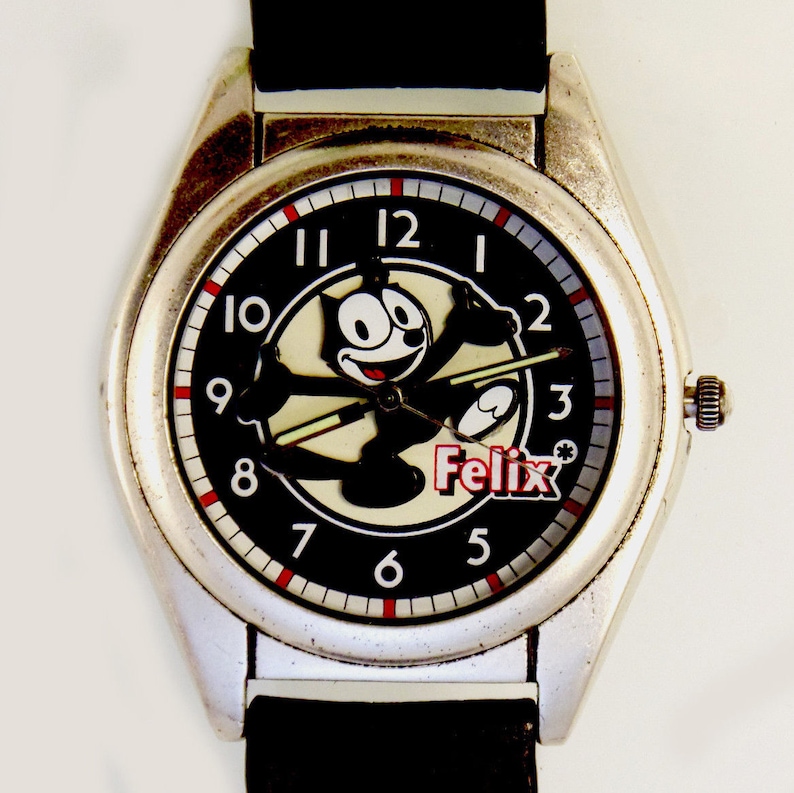 May include: A silver wristwatch with a black leather band. The watch face features a black background with white numbers and red accents. A cartoon image of Felix the Cat is in the center of the watch face. The text "Felix" is printed in red below the image.