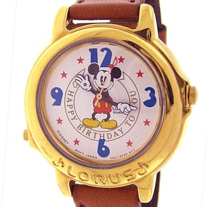 May include: A gold-tone wristwatch with a white face featuring Mickey Mouse and the text "Happy Birthday To You". The watch has a brown leather band.