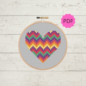 Puede incluir: Un patrón de punto de cruz de corazón de chevron colorido en un aro de bordado de madera. El patrón está en tonos de rojo, naranja, amarillo, verde, morado y rosa. El archivo PDF está disponible para descargar.