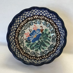 May include: A blue and white ceramic bowl with a scalloped edge. The bowl features a floral design with green leaves, blue flowers, and pink accents. The bowl has a brown and white geometric pattern around the rim.