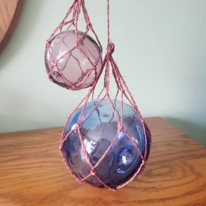 Puede incluir: Dos flotadores de pesca de vidrio, uno lavanda y otro azul, suspendidos en una red de cuerda anudada. Los flotadores son redondos y tienen una superficie texturizada. La cuerda es de color rosa apagado. Los flotadores cuelgan frente a una superficie de madera.