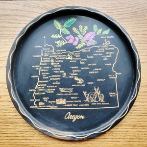 May include: A black serving tray with a gold rim and an Oregon map design. The map features cities, landmarks, and illustrations of wildlife. The word "Oregon" is written in a cursive font at the bottom.