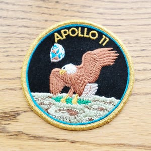Peut inclure: Un écusson brodé circulaire représentant un aigle brun atterrissant sur une surface lunaire grise sur fond noir. Le texte APOLLO 11 est brodé en jaune en haut, avec un petit graphique bleu et blanc de la Terre visible au-dessus de l'aigle.