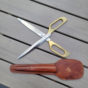 May include: Large silver and gold scissors with a brown leather sheath. The scissors have gold handles and a silver blade. The sheath is brown with a gold monogram. The scissors are designed for cutting fabric or other materials.