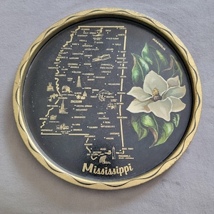 May include: Round black tray with a gold rim featuring a map of Mississippi. The tray includes a white magnolia flower and the word "Mississippi" in gold lettering. The tray is approximately 10 inches in diameter.