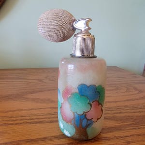 Puede incluir: Un frasco de perfume vintage de vidrio con un atomizador plateado y una bomba de tela beige. El frasco tiene un fondo blanco con un diseño pintado a mano de un árbol verde, azul y rosa con detalles dorados.