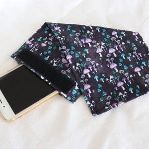 Op de afbeelding: Een zwarte telefoonhoes met een paddenstoelprint. De hoes heeft een klittenbandsluiting en is ontworpen om een smartphone te houden.