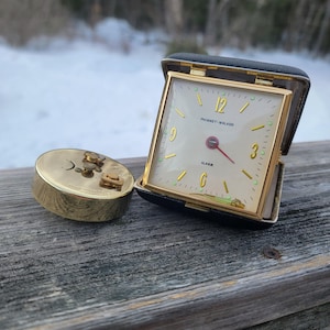 May include: A vintage gold-toned travel alarm clock with a square face, open in its black leather case. The clock face is cream-colored with gold numerals and hands. A separate gold-toned, cylindrical case is next to the clock.