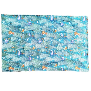 Op de afbeelding: Een rechthoekige, gewatteerde deken met een turquoise achtergrond en een herhalend patroon van witte en groene bomen, oranje vossen en blauwe en paarse wezens. De deken heeft een gestikte rand.