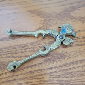 May include: Antique brass nutcracker with a bird-like design. The nutcracker features a detailed head with a blue accent and curved handles. The brass has a worn, vintage appearance, suggesting age and use. A decorative kitchen tool.