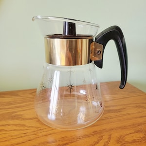 May include: A vintage glass coffee carafe with a gold-colored metal band and a black handle. The clear glass carafe features a starburst pattern. The carafe is sitting on a wooden surface.