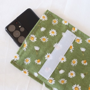 Könnte beinhalten: Eine grüne Stoff-Handyhülle mit einem weißen Gänseblümchenmuster und einem weißen Klettverschluss. Ein schwarzes Smartphone mit einem Kameraobjektiv befindet sich in der Hülle.