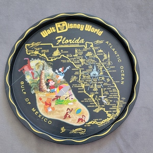 May include: Black metal serving tray with a gold rim and a map of Florida featuring Walt Disney World. The tray includes illustrations of Disney characters like Mickey Mouse, Donald Duck, and Winnie the Pooh. The tray is approximately 12 inches in diameter.