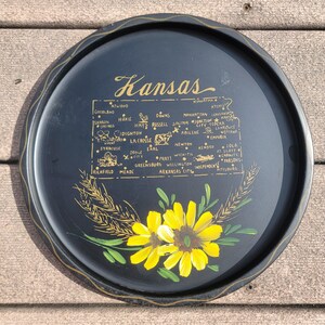 May include: A round, black serving tray with a gold rim and a painted map of Kansas. The tray features the word "Kansas" in gold script, along with yellow sunflowers and wheat stalks. The tray measures approximately 12 inches in diameter.