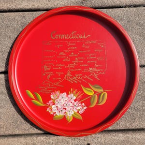 Peut inclure: Un plateau rond en métal rouge avec un bord doré et une carte peinte du Connecticut. La carte comprend des noms de villes en lettres dorées, avec un motif floral dans la partie inférieure. Le mot "Connecticut" est écrit en lettres dorées.