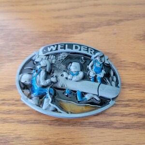 Può includere: Fibbia ovale in metallo con tre figure di saldatura. La parola "WELDER" è in alto. Le figure indossano tute blu e caschi. La fibbia ha una finitura argento e nera.