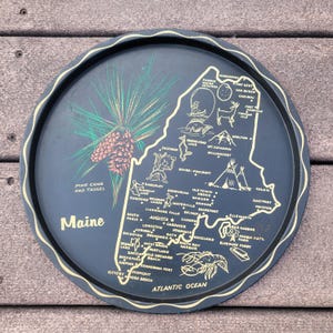 May include: A round, dark blue tray with a gold rim and a map of Maine. The tray features illustrations of a pine cone, a lobster, and various landmarks. Text includes "Maine", "Pine Cone and Tassel", and city names.
