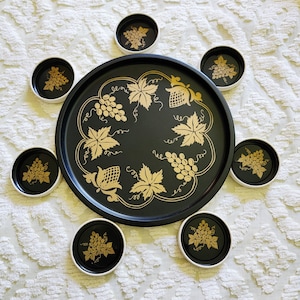 May include: A large round black serving tray surrounded by six smaller matching round coasters. Each piece features a gold-colored graphic design of grape clusters and vine leaves. The tray and coasters have a smooth black finish with gold accents.