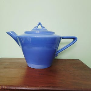 May include: A blue ceramic teapot with a lid and handle. The teapot has a spout and a handle. The lid has a triangular handle. The teapot is sitting on a wooden surface. The teapot is a solid blue color.