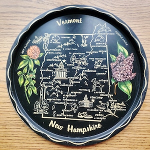 May include: A black metal tray featuring a map of Vermont and New Hampshire, with gold trim. The tray includes illustrations of landmarks, flowers, and text. The words "Vermont" and "New Hampshire" are prominently displayed.