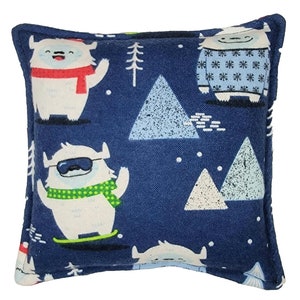 May include: A blue square pillow with a white yeti wearing a red scarf and a green scarf, a blue sweater with white snowflakes, and a blue and white mountain pattern.