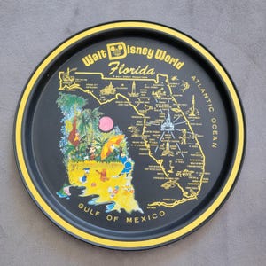 May include: A black and yellow round tray featuring a map of Florida with Walt Disney World landmarks. The tray has a yellow border and includes illustrations of Disney characters and the text "Walt Disney World Florida".