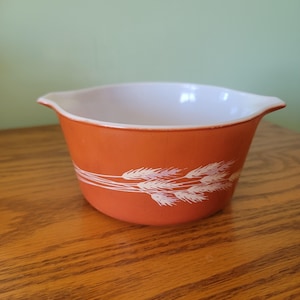 Puede incluir: Un cuenco Pyrex naranja con interior blanco y un diseño de trigo. El cuenco tiene forma redondeada con borde acampanado y dos asas pequeñas. El diseño de trigo es blanco con sutiles acentos lavanda.