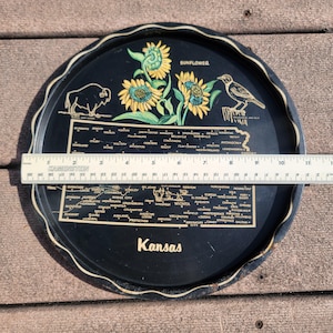May include: A black, round tray with a white scalloped edge. The tray features a map of Kansas, sunflowers, a bison, and a bird. The word "Sunflower" is above the flowers, and "Kansas" is at the bottom. The tray measures approximately 10 inches in diameter.