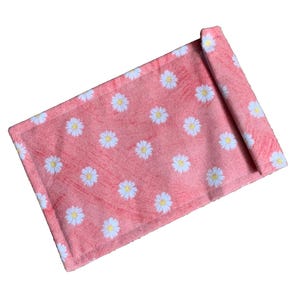 Op de afbeelding: Een koraalkleurige, rechthoekige placemat met een herhalend patroon van witte madeliefjes met gele centra. De placemat is aan één uiteinde opgerold, wat het bloemmotief laat zien. De randen zijn netjes gestikt.