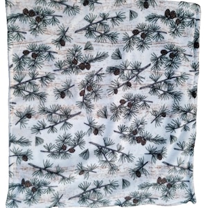 May include: White fabric featuring a repeating pattern of green pine branches and brown pine cones. The background has a light brown, wood-grain texture. The fabric's edges are slightly frayed.