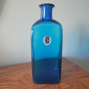 May include: A tall, rectangular blue glass bottle with a rounded neck and a small, circular label. The bottle has a gradient of blue hues, with a darker shade at the top and bottom. The label features text and a circular design. The bottle is displayed on a wooden surface.