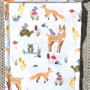 May include: White fabric pouch with a whimsical woodland scene. The design features foxes, deer, turtles, mice, mushrooms, and frogs. The pouch has a fold-over flap and is likely used for storage or carrying small items.