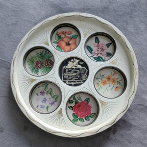 Puede incluir: Una bandeja redonda y blanca con ribete dorado y un diseño floral decorativo. La bandeja presenta siete insertos circulares con imágenes de flores y un mapa del estado de Pensilvania. La bandeja es de metal.