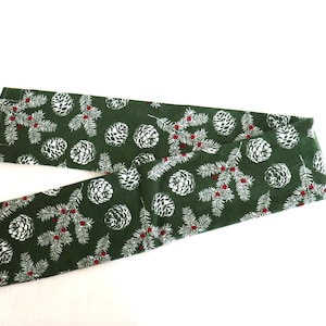 May include: A green fabric strip with a repeating pattern of white pine cones and red berries on green pine branches.