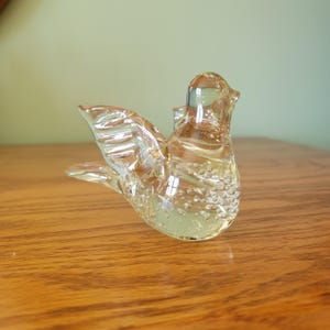 Könnte beinhalten: Eine klare Glasvogel-Figur mit gelblichem Farbton. Der Vogel hat ausgebreitete Flügel und einen detaillierten Körper mit kleinen Luftblasen. Die Figur steht auf einer Holzoberfläche.