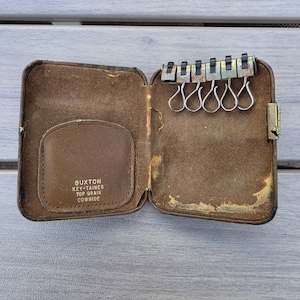 May include: A brown leather key case with a metal frame and six silver-toned key hooks. The interior features a pocket with the embossed text BUXTON KEY-TAINER TOP GRAIN COWHIDE. The case is open, revealing the worn brown suede-like lining.