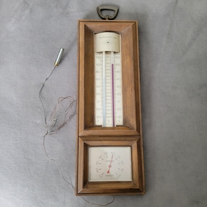 Puede incluir: Un termómetro e higrómetro de madera con un abridor de botellas de metal en la parte superior. El termómetro muestra la temperatura en grados Fahrenheit, y el higrómetro muestra los niveles de humedad. El marco es de color marrón cálido.