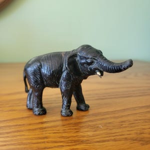 May include: A small, dark brown elephant figurine with detailed texture. The elephant has a long trunk, large ears, and visible tusks. The figurine is standing on a wooden surface.