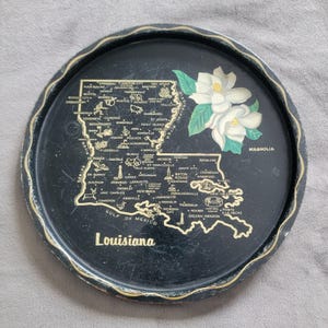 May include: Black, round serving tray featuring a map of Louisiana in gold, with city names. The tray has a gold rim and a painted magnolia flower design. The word "Louisiana" is printed in gold.