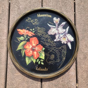 May include: Round black tray with a gold rim featuring a map of the Hawaiian Islands. The tray is decorated with illustrations of hibiscus and orchid flowers. The words "Hawaiian" and "Islands" are printed on the tray.