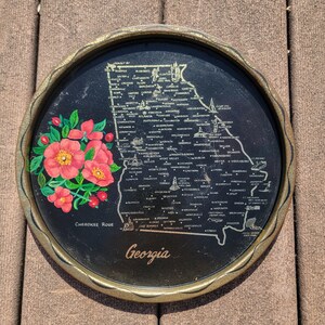 May include: Round black tray with a gold rim featuring a map of Georgia. The tray has a floral design with pink flowers and the words "Cherokee Rose" and "Georgia" in white script. The tray is approximately 10 inches in diameter.