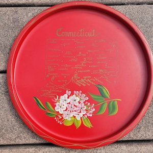 Może przedstawiać: Okrągły, czerwony talerz do serwowania ze złotą obwódką i złotą mapą stanu Connecticut. Słowo "Connecticut" jest napisane złotymi literami. Na tacy namalowano kwiatowy wzór z białymi i różowymi kwiatami oraz zielonymi liśćmi.